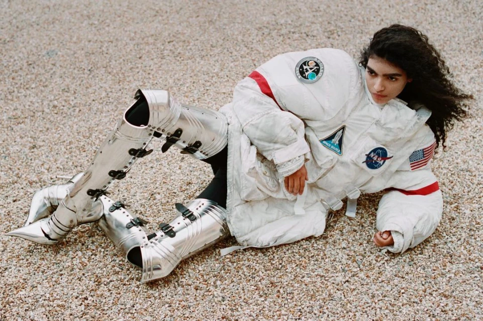 Woman wearing a spacesuit and medieval armor poses in a ground-level shot. The outdoor setting features neutral-toned gravel.