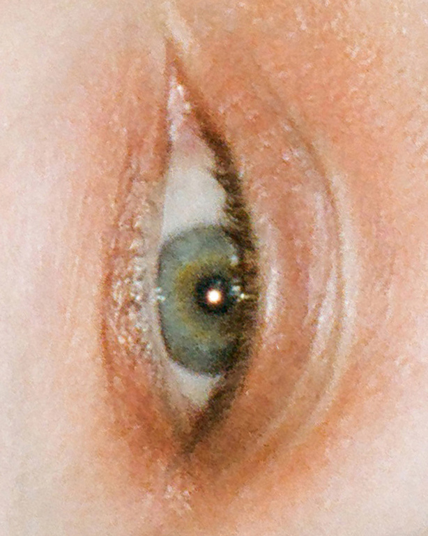 Close-up photo of an eye with green, grey, and gold tones, and visible eyelashes and skin texture. Intimate beauty portrait.