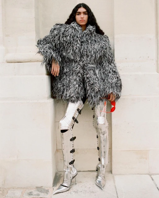 A woman in a feathered coat and silver boots poses against a white wall in a fashion shot with neutral tones and stylish attire.