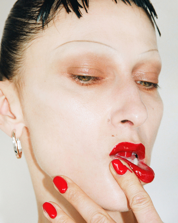 Close-up portrait of a model with red lipstick and nail polish, finger to lips, wet-look hair, pale skin, silver earring, and white background.