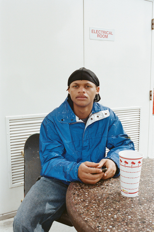 Young man in a blue jacket sits at an outdoor table with a drink. He is wearing a black head covering, and is seated next to a skateboard.