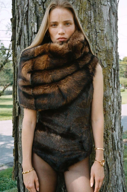 Adult female fashion model wearing a fur bodysuit and shawl posing outdoors next to a large tree, with dark leggings and gold bracelets.