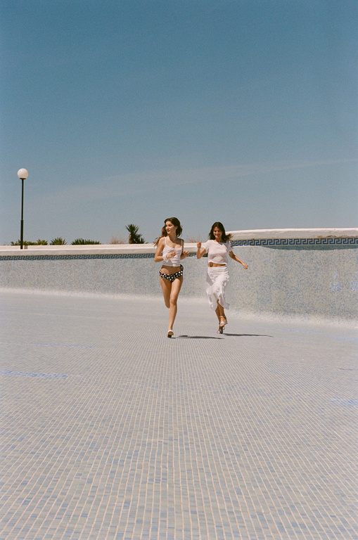 Two women in swimwear run along the bottom of a drained swimming pool under a clear sky, enjoying the bright sunny day.