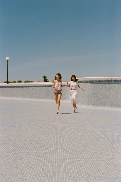 Two women in swimwear run along the bottom of a drained swimming pool under a clear sky, enjoying the bright sunny day.