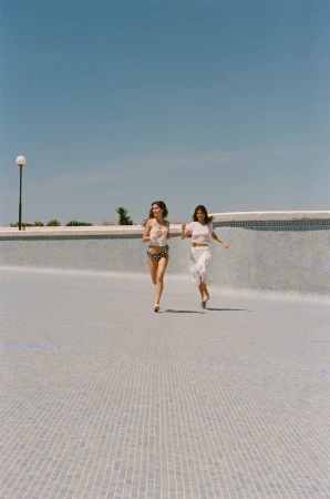 Two women in swimwear run along the bottom of a drained swimming pool under a clear sky, enjoying the bright sunny day.