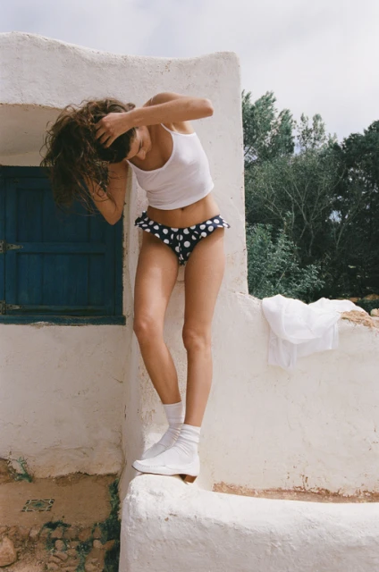 Woman poses in swimwear and socks against a white stucco wall with a blue window, showcasing fashion and outdoor summer style.