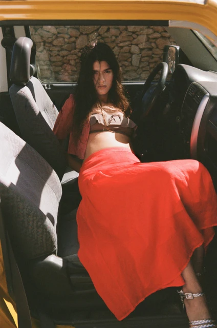 A woman with long dark hair poses in the driver's seat of a convertible, wearing a brown top and red skirt, with a stone wall in the background.