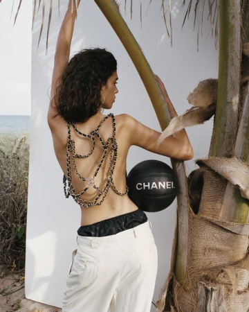 Woman in white trousers holding a black Chanel basketball while standing outdoors by a palm tree and white wall