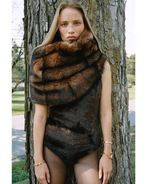A fair-skinned woman poses outdoors in a brown fur bodysuit, accessorized with gold bracelets, next to a tall, textured tree trunk.