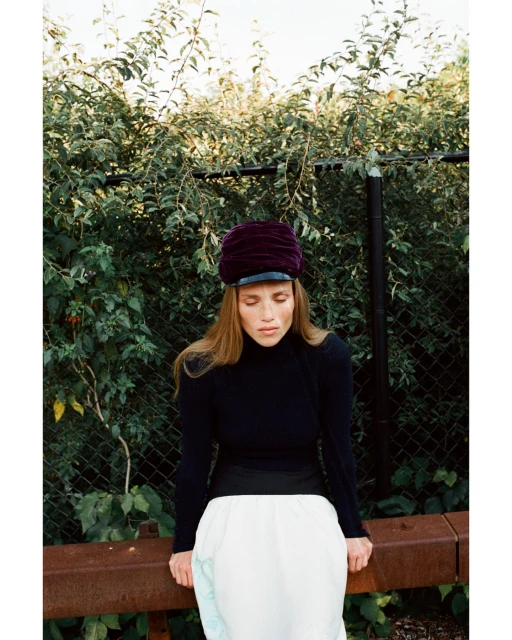 Woman in a purple cap and black turtleneck poses outdoors with green foliage in the background. She leans against a rusty railing.