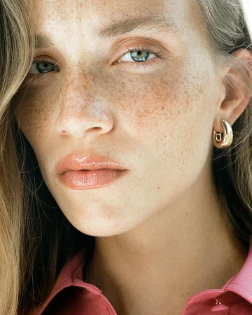 Close-up portrait of a fair-skinned adult female with freckles, wearing a pink top and gold hoop earrings, showcasing beauty.
