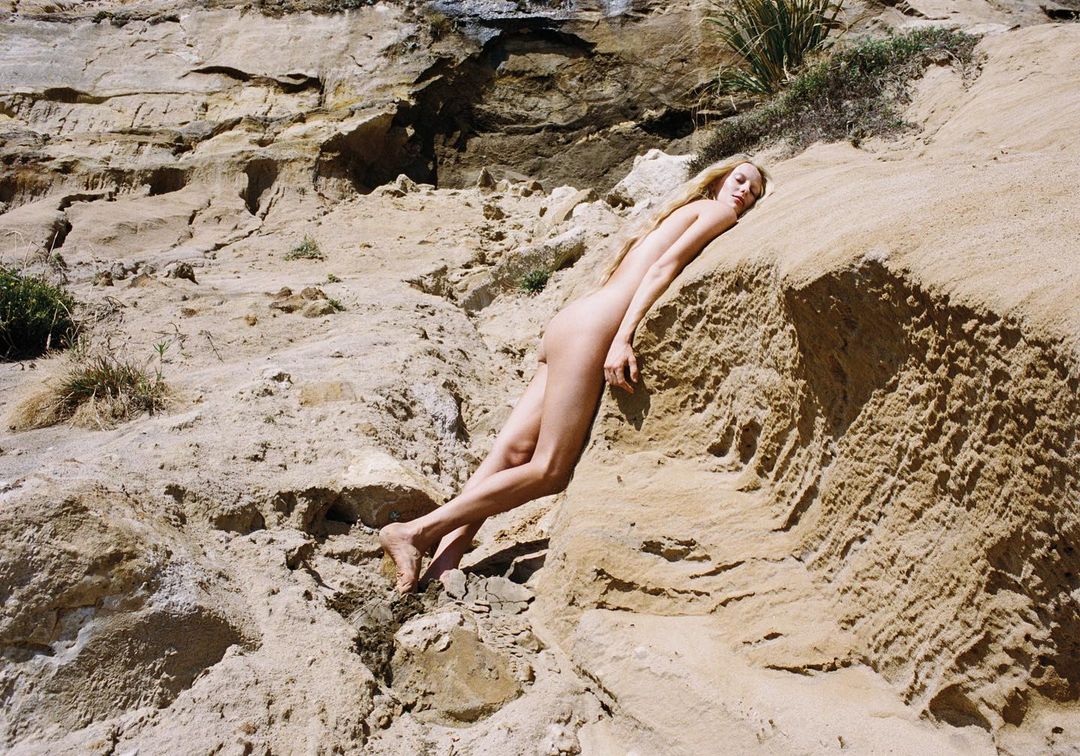 Nude adult woman with long blonde hair reclines on a textured tan rock formation near a cliff face in an outdoor, natural setting.