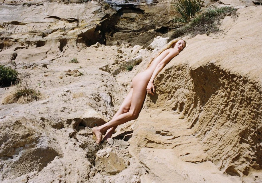 Nude adult woman with long blonde hair reclines on a textured tan rock formation near a cliff face in an outdoor, natural setting.