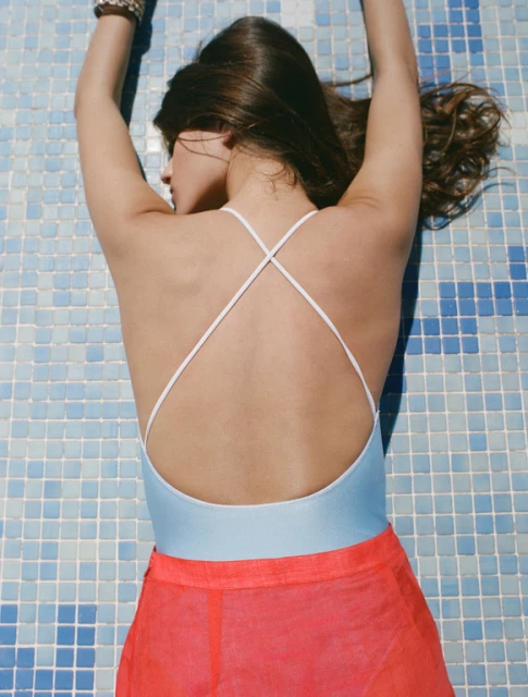 Young woman lying on blue tiled surface wearing a light blue swimsuit with thin white straps and an orange sheer cover-up skirt.