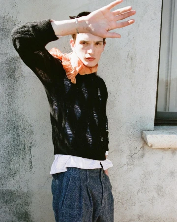Young man raising hand to block sun while wearing black knit sweater and peach ruffled scarf outdoors