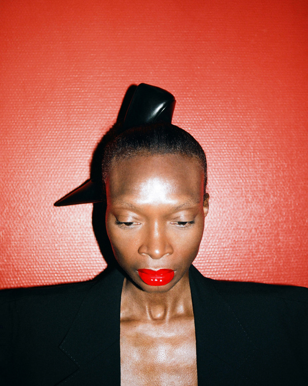 Portrait of a Black woman with red lipstick and avant-garde hat against a textured red backdrop wearing a black jacket. Fashion close-up.