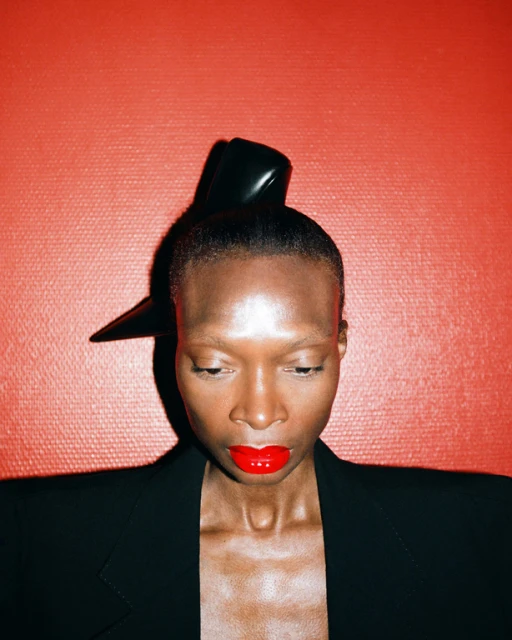 Portrait of a Black woman with red lipstick and avant-garde hat against a textured red backdrop wearing a black jacket. Fashion close-up.