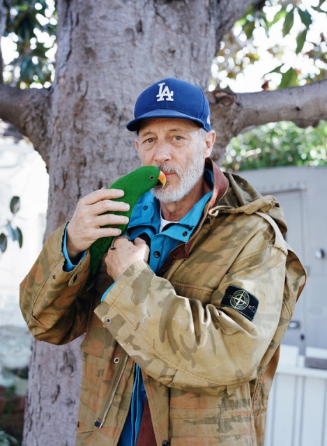 Adult male poses outdoors with a green parrot. He wears a Los Angeles Dodgers baseball cap and camouflage jacket by a tree.