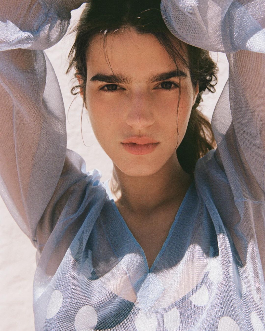 Close-up portrait of a young woman with light brown hair and natural makeup, wearing a light blue, polka-dot blouse, with hands above her head.