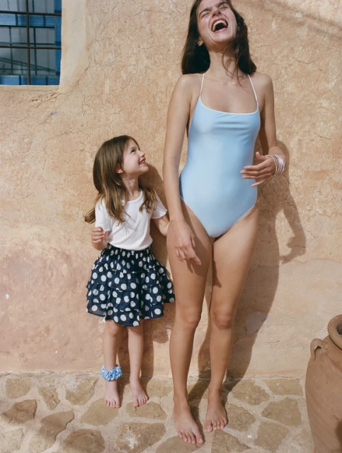 Woman in a swimsuit with a young girl in a polka dot skirt laughing in front of a tan stucco wall with stone base and an earthenware pot.