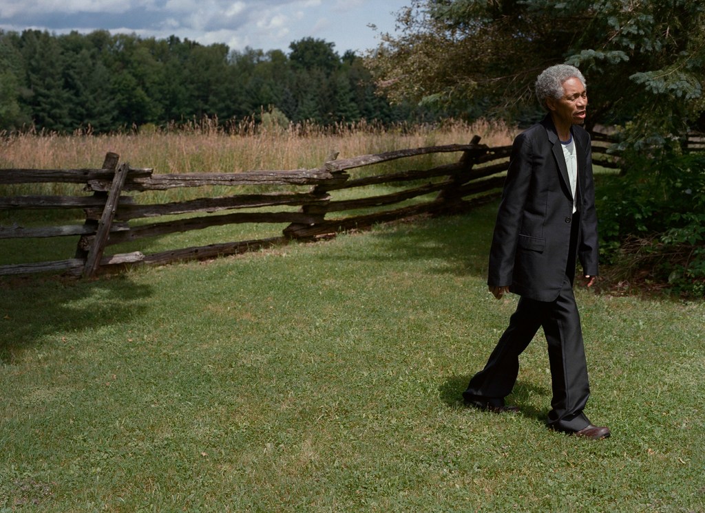Mature woman in a black suit walks through a green field near a wooden fence on a sunny day with trees in the background.