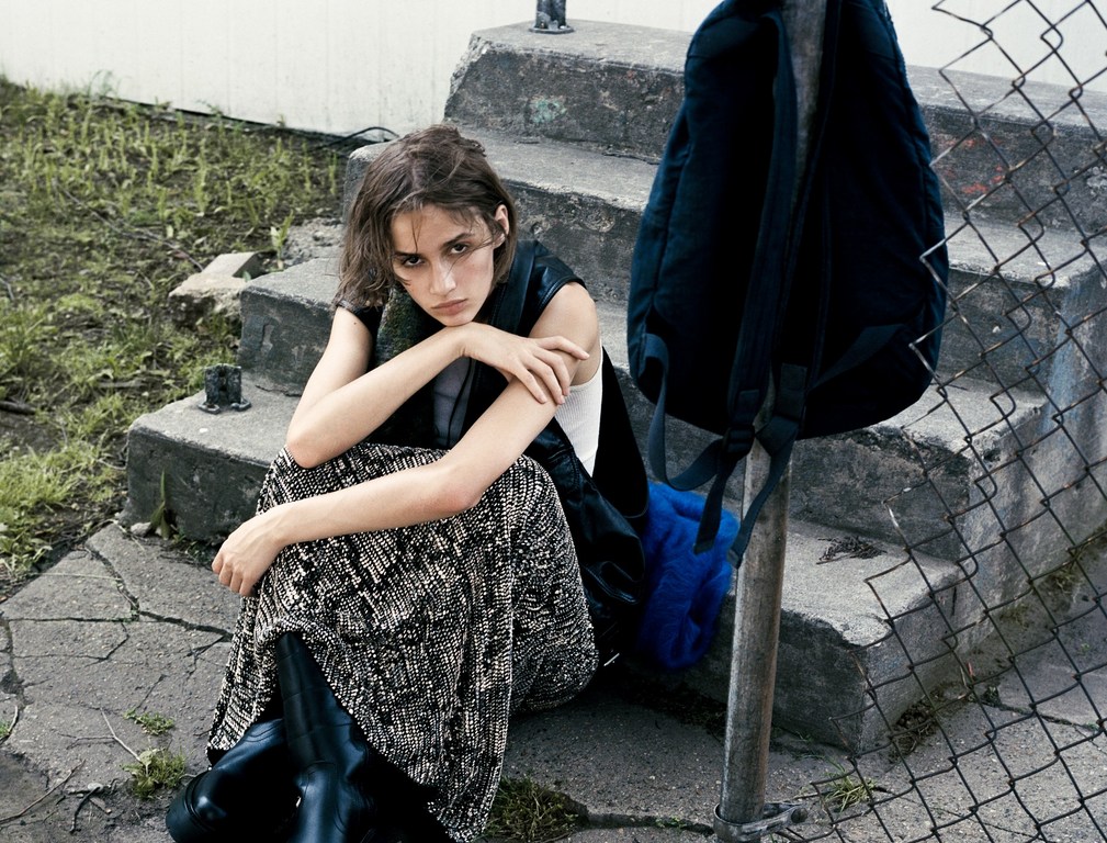 Young woman in a patterned skirt and dark boots seated on concrete steps next to a chain-link fence by photographer Thistle Brown.