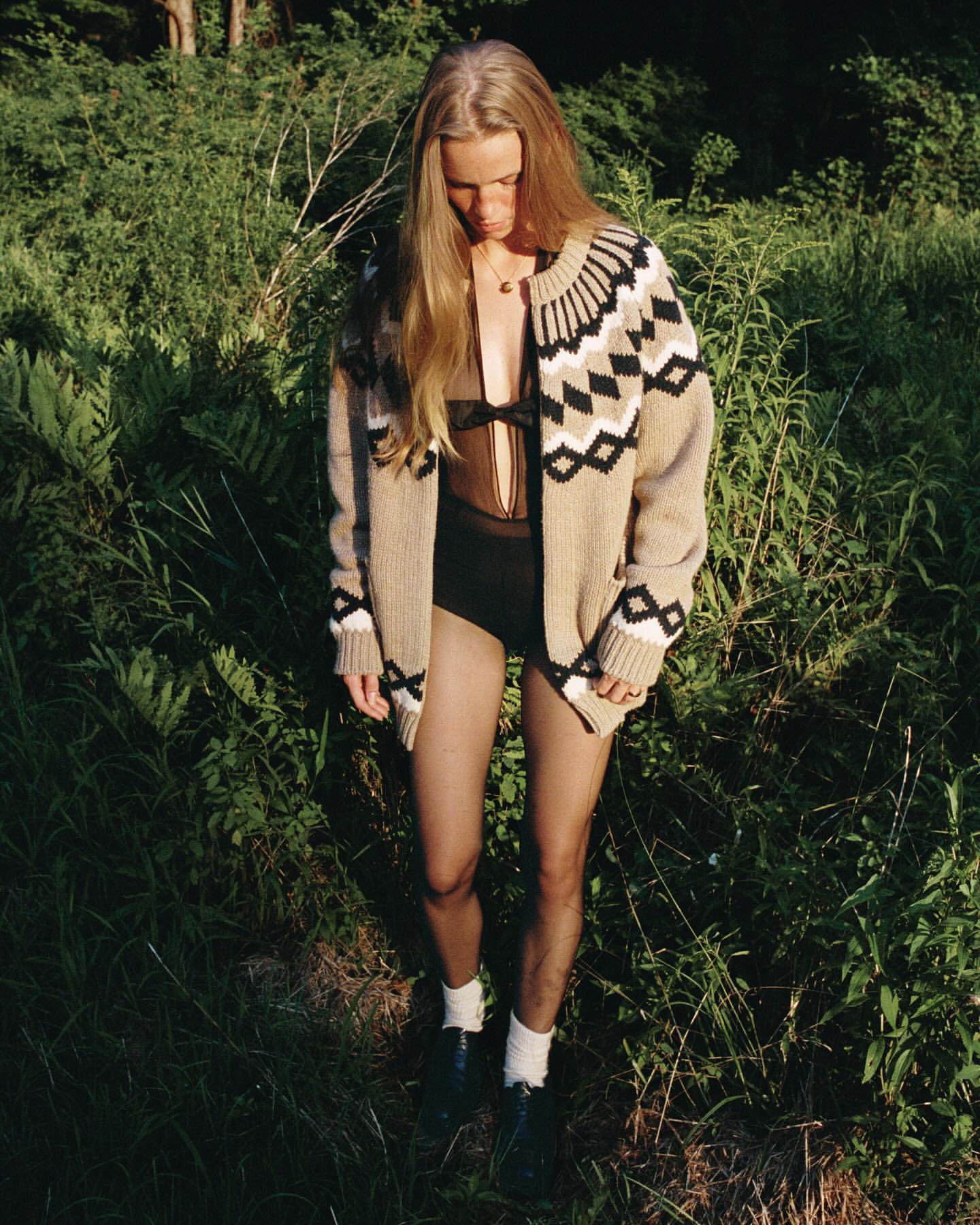 A blonde-haired woman in a cardigan and dark leotard stands in a grassy, wooded area, looking down with soft, natural light.