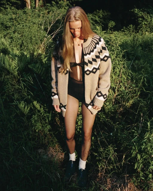 A blonde-haired woman in a cardigan and dark leotard stands in a grassy, wooded area, looking down with soft, natural light.