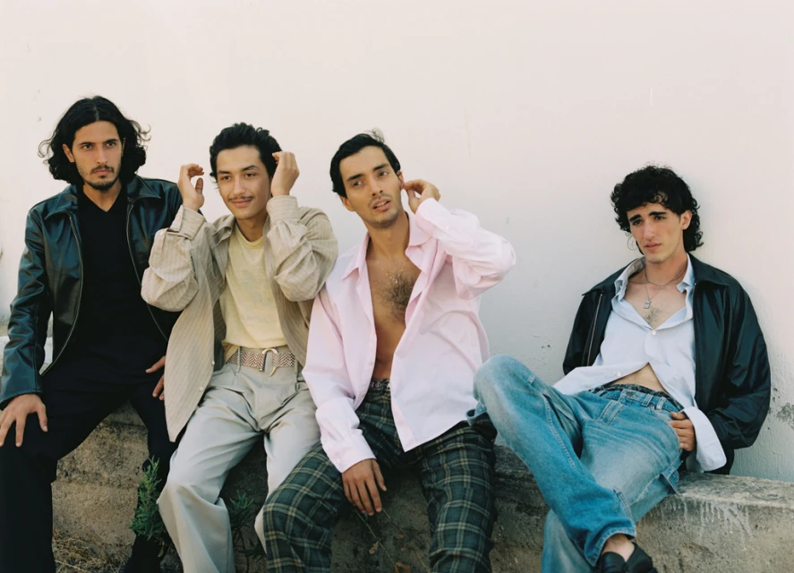 Four stylish young men sit together in front of a white wall, wearing fashionable shirts, jackets, and jeans in a casual outdoor setting.