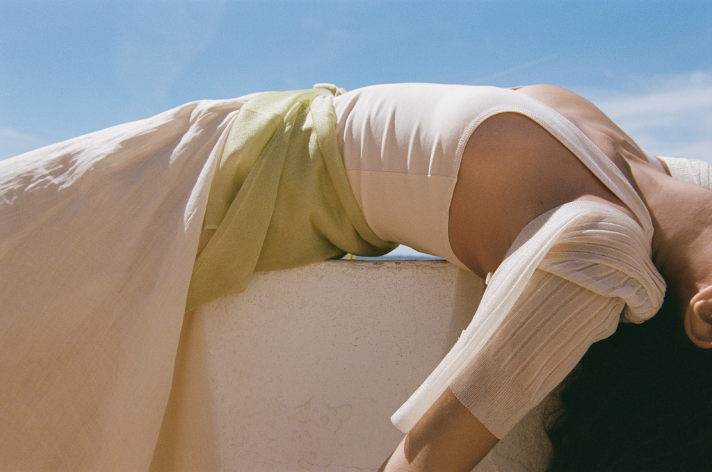 Adult female in light-colored dress leans back against a low white wall under a bright blue sky, captured in a relaxed, stylish outdoor setting.