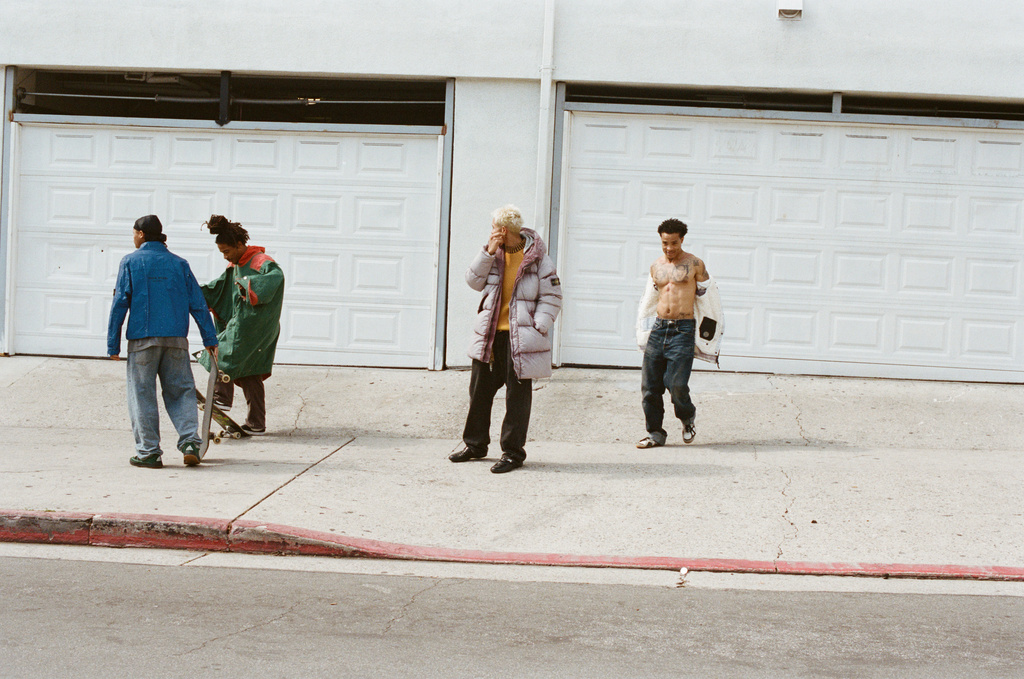 Four people stand outdoors in casual streetwear near garage doors on a sunny day. One person holds a skateboard, another walks shirtless.