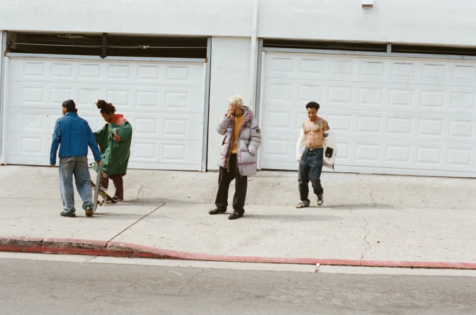 Four people stand outdoors in casual streetwear near garage doors on a sunny day. One person holds a skateboard, another walks shirtless.