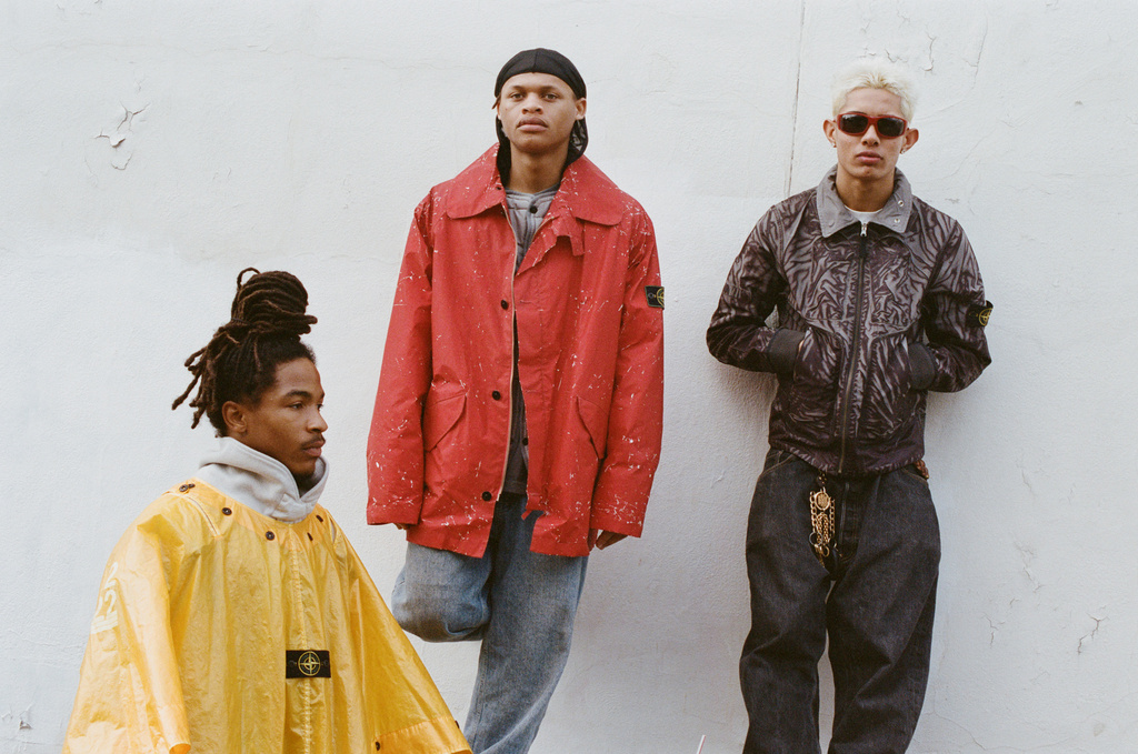 Three men pose against a light wall wearing casual, colorful Stone Island jackets and jeans in an outdoor, eye-level, medium shot.