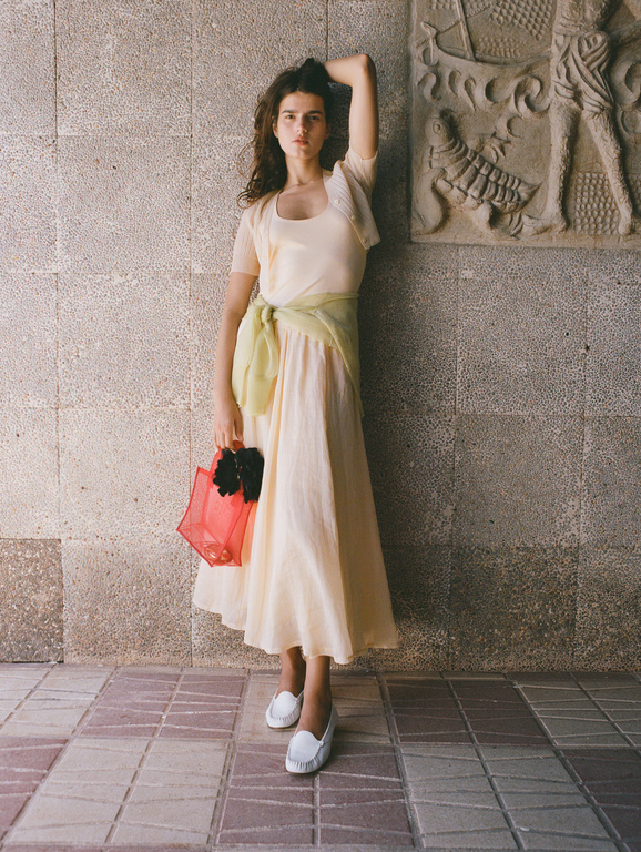 Woman in a dress leans against a stone wall with bas-relief carvings, holding a red tote bag. She wears white shoes and a light green shawl.