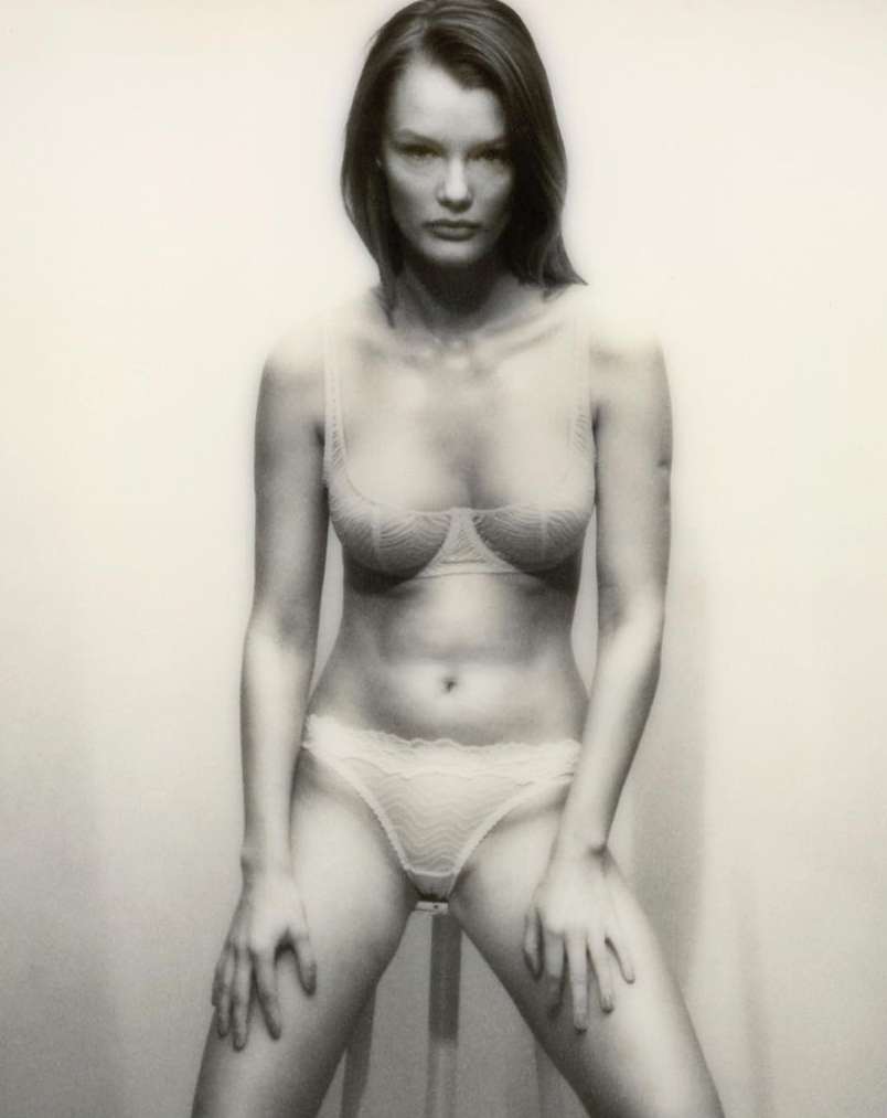 Artistic studio portrait of a woman in lingerie, posing with hands on a stool, soft lighting, and neutral tones.