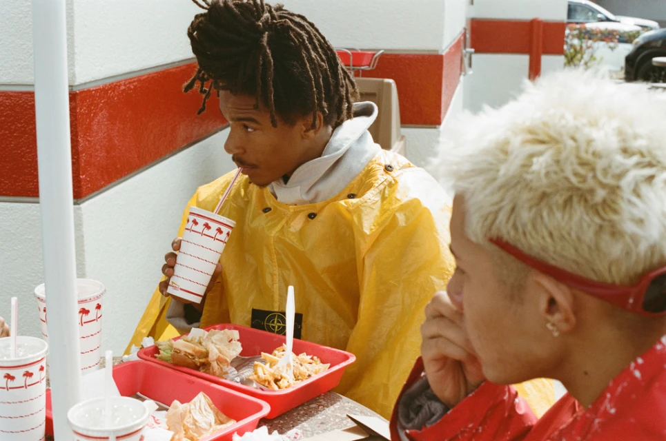 Two people sit at an outdoor table at In-N-Out Burger, enjoying food and drinks under bright daylight. One wears a yellow jacket.