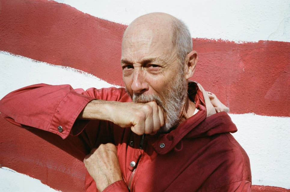 Close-up of a bald adult man in a red jacket standing in front of a red-and-white striped wall, with his fist near his mouth.