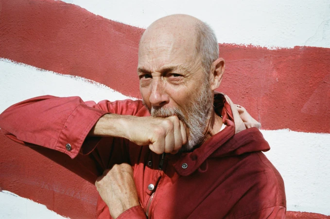 Close-up of a bald adult man in a red jacket standing in front of a red-and-white striped wall, with his fist near his mouth.