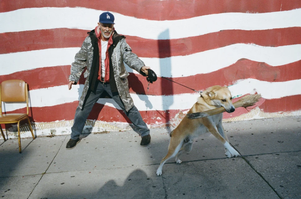 Man in a metallic coat walks a dog that is carrying a broom in its mouth in front of a red and white striped wall.