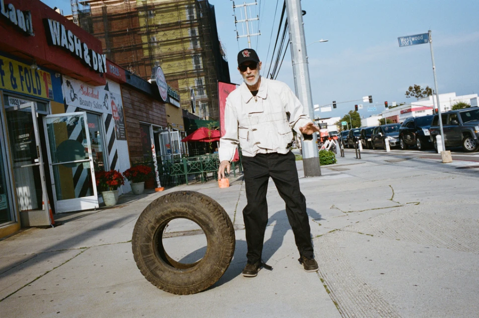 Adult male stands on a sidewalk next to a large tire in an urban setting with buildings and traffic in the background on a sunny day.