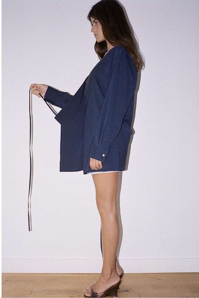 A young woman stands in profile, wearing a large blue button-down shirt and clear high-heeled sandals, holding a striped sash.