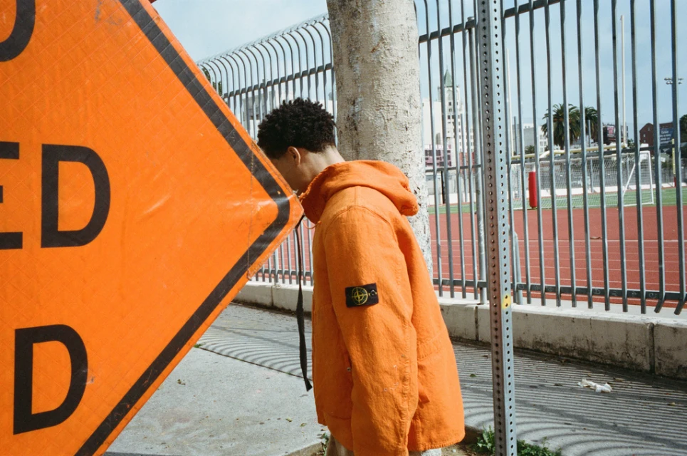 Adult male in orange jacket poses next to construction sign and metal fence near urban sports field on a sunny day.