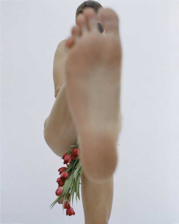 Close-up shot of a nude adult with a bouquet of red tulips at the waist, looking into the camera with a blurred foot.