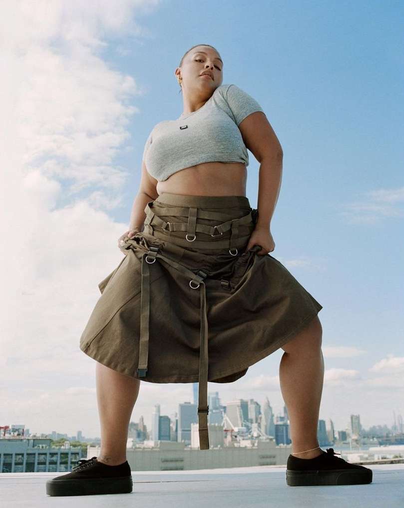Woman in a crop top and skirt stands on a rooftop with a cityscape in the distance under a cloudy sky, shot from a low angle.