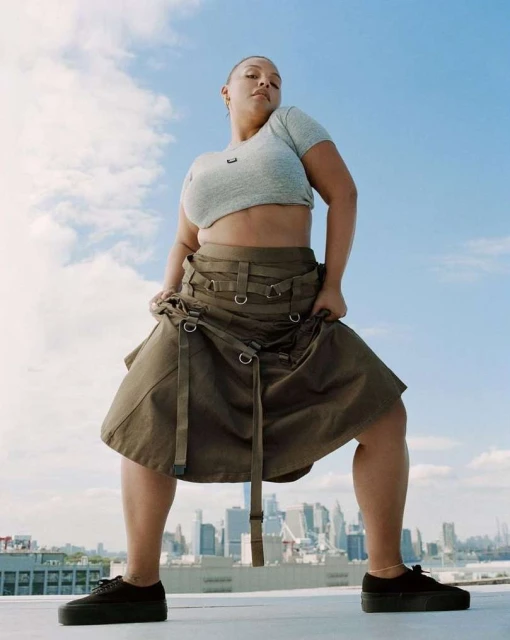 Woman in a crop top and skirt stands on a rooftop with a cityscape in the distance under a cloudy sky, shot from a low angle.