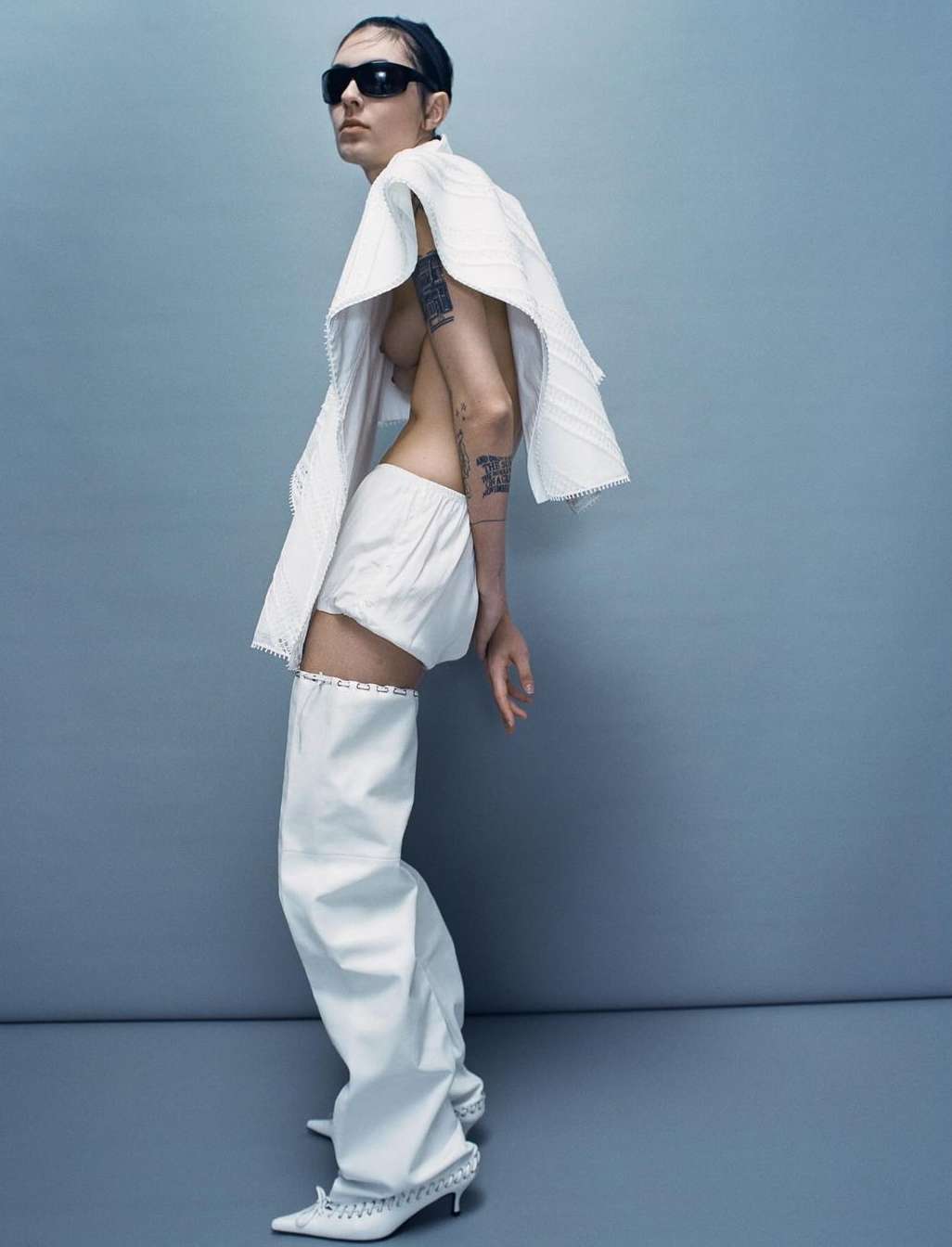 A fashion photo shows a slim adult female posing in white shorts, a white shawl-like top, white leg coverings and black sunglasses, indoors.