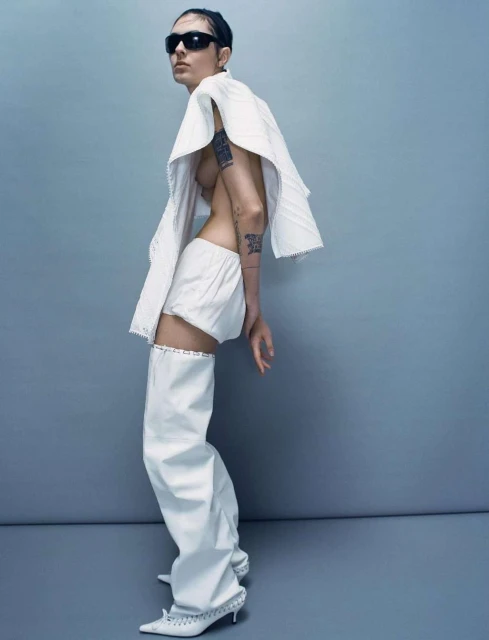 A fashion photo shows a slim adult female posing in white shorts, a white shawl-like top, white leg coverings and black sunglasses, indoors.