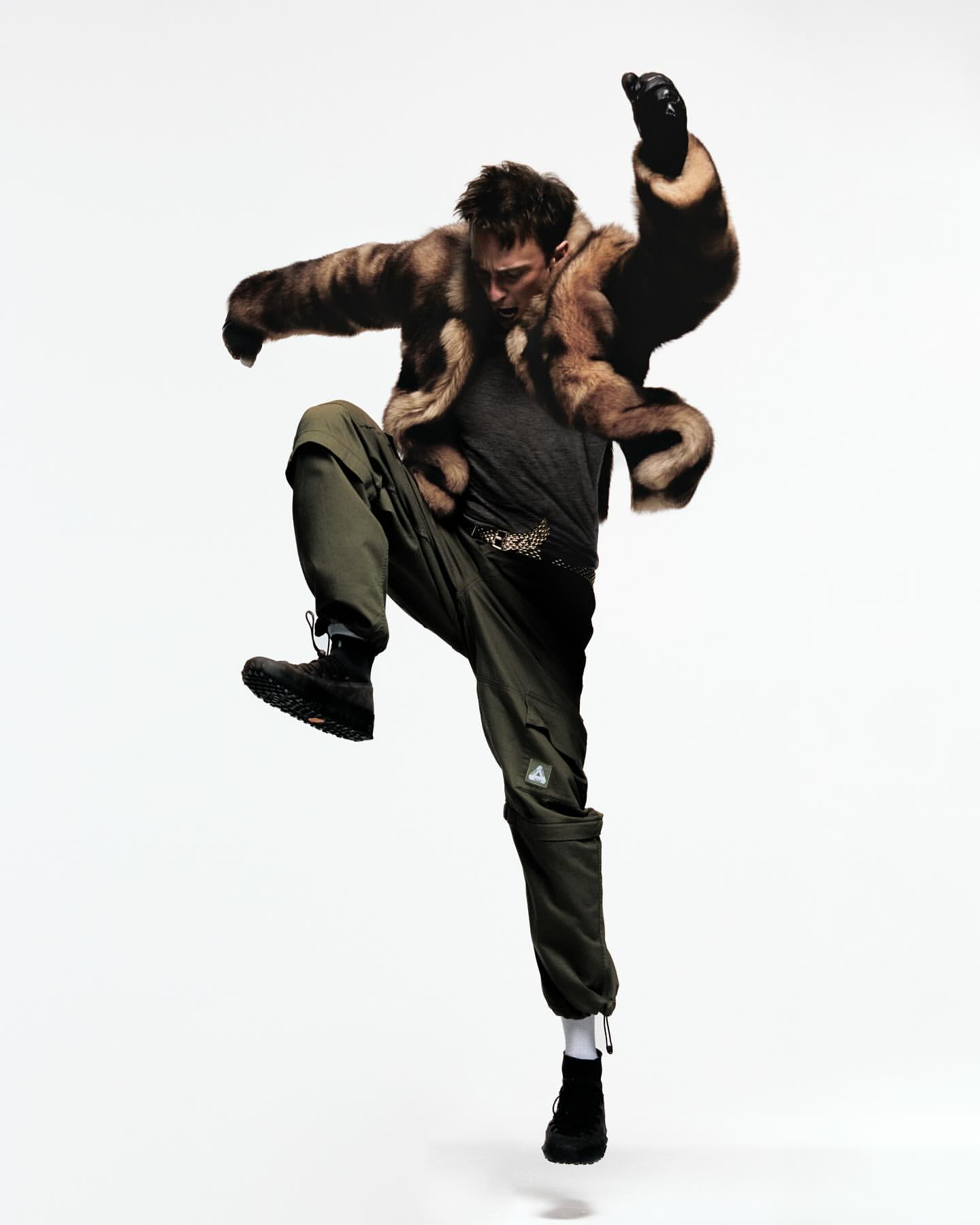A male fashion model strikes a dynamic pose mid-air in a fur coat, cargo pants and black boots set against a clean, white backdrop.