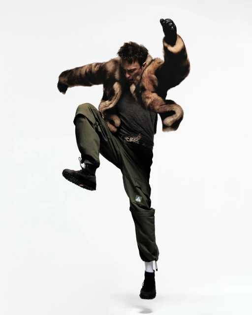A male fashion model strikes a dynamic pose mid-air in a fur coat, cargo pants and black boots set against a clean, white backdrop.