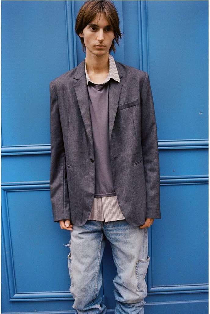 Young male model in grey blazer, striped shirt, and light denim jeans standing against a vibrant blue paneled door.